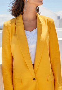 Geel blazer met een gladde textuur, met inkepingen op de revers en een enkele knoopsluiting, gecombineerd met een witte top met een gestructureerd patroon.