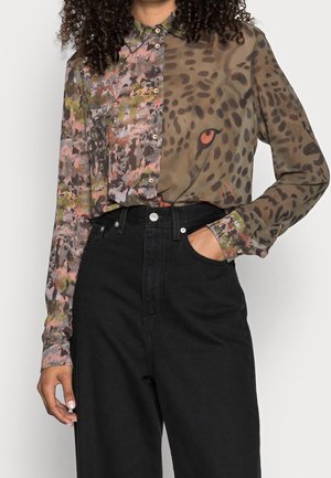 Frau trägt ein Hemd mit Knöpfen, das halb mit Tarn- und halb mit Leoparden-Gesichtsprint bedruckt ist, in eine hoch taillierte schwarze Hose gesteckt.