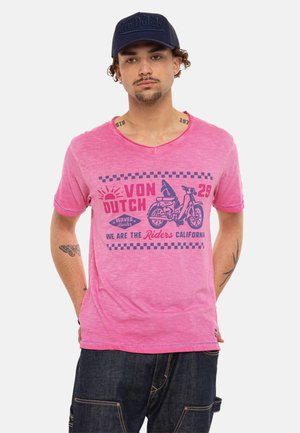 Homme portant un t-shirt rose Von Dutch avec un motif de moto et une casquette bleu foncé, debout les mains dans le dos sur un fond uni.