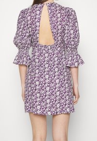 Robe violette avec des motifs floraux blancs, manches courtes et dos ouvert. Dotée d'un design de manches évasées et d'une taille ajustée.