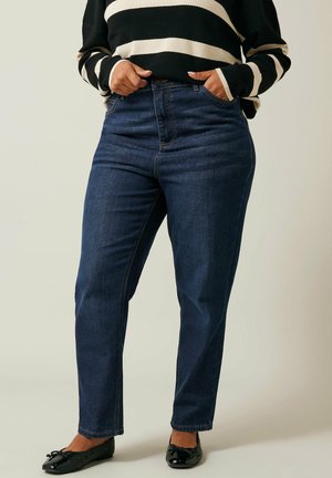 Jeans a vita alta blu scuro con una vestibilità slim, dotati di tasche e dettagli di cucitura sottili. Indossati con un maglione a righe nero e crema.