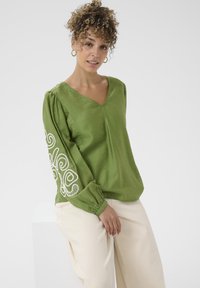 Blouse verte légère avec col en V, manches bouffantes et motifs tourbillonnants brodés en blanc sur le bras gauche. Associée à un pantalon beige.