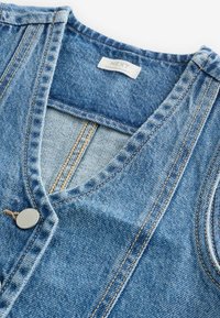 Gilet senza maniche in denim blu con scollo a V, cuciture color cuoio, bottone in metallo e etichetta di tessuto con la scritta "NEXT Made in Bangladesh" all'interno del colletto.