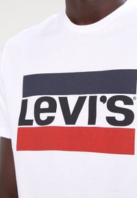 Vit bomulls T-shirt med en stor svart "Levi's"-logotyp, med en blå och röd rektangulär bakgrund ovanför och under texten.