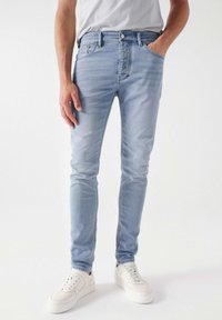 Vaqueros de denim azul claro con un corte ajustado, que cuentan con bolsillos frontales, costuras en contraste y una textura ligeramente desgastada. Combinados con zapatillas blancas.