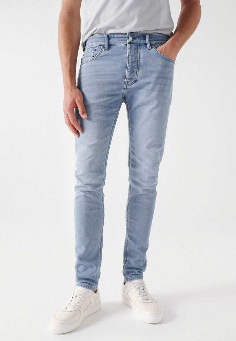 Vaqueros de denim azul claro con un corte ajustado, que cuentan con bolsillos frontales, costuras en contraste y una textura ligeramente desgastada. Combinados con zapatillas blancas.