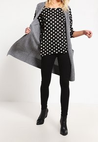 Schwarz-creamfarbene gepunktete Bluse, graue offene Strickjacke, schwarze Leggings und Ankle Boots. Glatte Texturen, entspannter Schnitt, geschichtetes Design.
