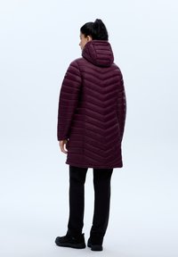 Lang, puffy kastanjebruine jas met een chevronpatroon, ritskap, en elastische manchetten, gecombineerd met zwarte broek en laarzen. Achteraanzicht weergegeven.