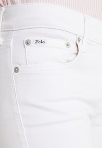 Bílé džíny z denimu s hladkou texturou, vybavené štítkem Polo, stříbrným kováním a standardním designem s pěti kapsami.