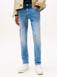 Férfi világoskék szűk fazonú farmerben, fehér tornacipőben, tengerészkék pulóverben és bézs dzsekiben, egyik kezét a zsebében tartva, fehér háttér előtt.