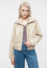 Mlada žena s dugom plavom kosom koja nosi bež jaknu preko ljubičaste majice i širokim plavim trapericama, stoji naslonjena na jednobojnu pozadinu.