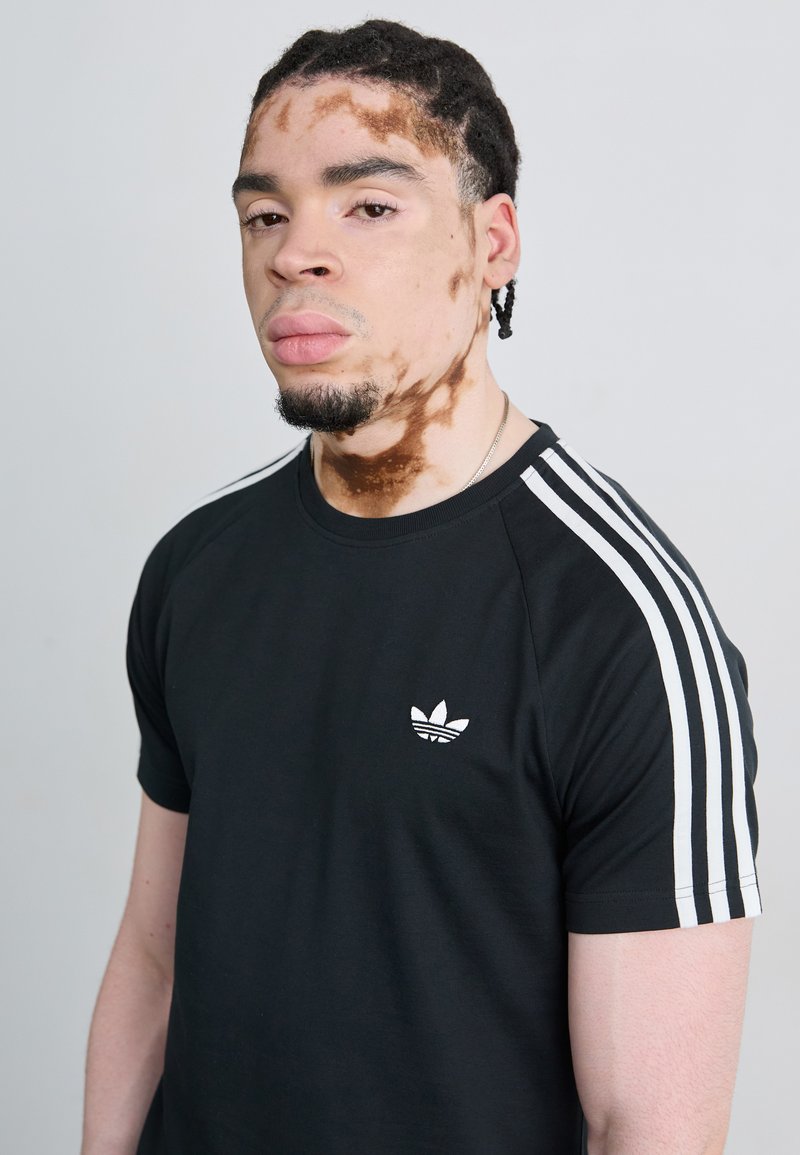 Schwarzes T-Shirt mit kurzen Ärmeln und weißen Streifenakzenten auf den Schultern sowie gesticktem Logo auf der Brust. Glattes Baumwollmaterial und regulärer Schnitt.