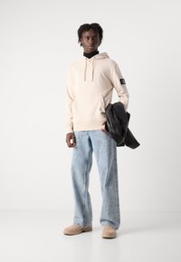 Felpa beige con cappuccio e tasca frontale, jeans leggermente larghi di colore blu chiaro, stivali color nocciola e una giacca di pelle nera appoggiata sul braccio.