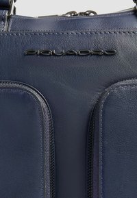 Borsa di pelle blu navy scuro con texture liscia, dotata di due tasche con cerniera e un logo "Piquadro" lucido impresso sulla parte anteriore.