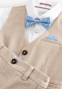 Bež prsluk i hlače od odijela s bijelom košuljom, svijetloplavom leptir mašnom s geometrijskim uzorkom i odgovarajućim džepnim maramicom.