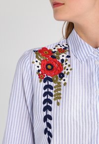 Camisa a rayas azul y blanca con intrincados bordados florales en rojo, azul marino y verde en el hombro. Cierre de botones y detalles en el cuello.