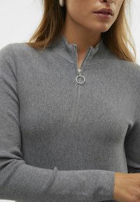 Grå zip-up tröja i texturerad tyg. Har en hög krage och en silverfärgad dragkedja med ett runt drag, långa åtsittande ärmar.