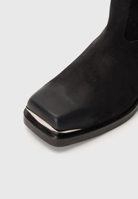 Stivale alla caviglia in suede nero con punta quadrata e design elegante. Caratterizzato da una punta lucida nera e un accento argento sul bordo della punta.