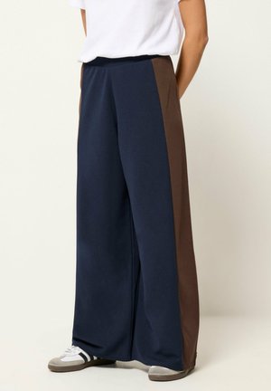 Persona con pantalones anchos azul marino con paneles laterales marrones, camisa blanca y zapatillas grises con rayas negras.