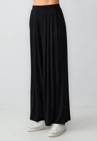 Pantalon noir à jambes larges en tissu doux, avec une taille haute et des plis, associé à des baskets blanches sur un fond clair.