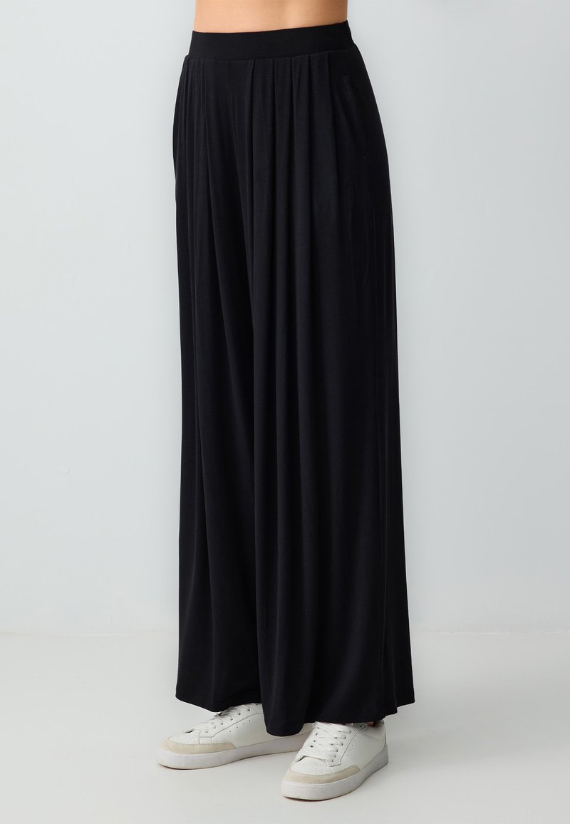 Pantalon noir à jambes larges en tissu doux, avec une taille haute et des plis, associé à des baskets blanches sur un fond clair.