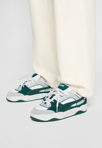 Sneaker atletiche bianche e verdi con una tomaia in suede testurizzato e mesh, lacci bianchi prominenti e una suola di gomma spessa.