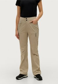 Beige outdoor broeken met een rechte pasvorm, voorzien van twee steekzakken en subtiele ritsen. Draagt een zwart shirt en zwarte schoenen.