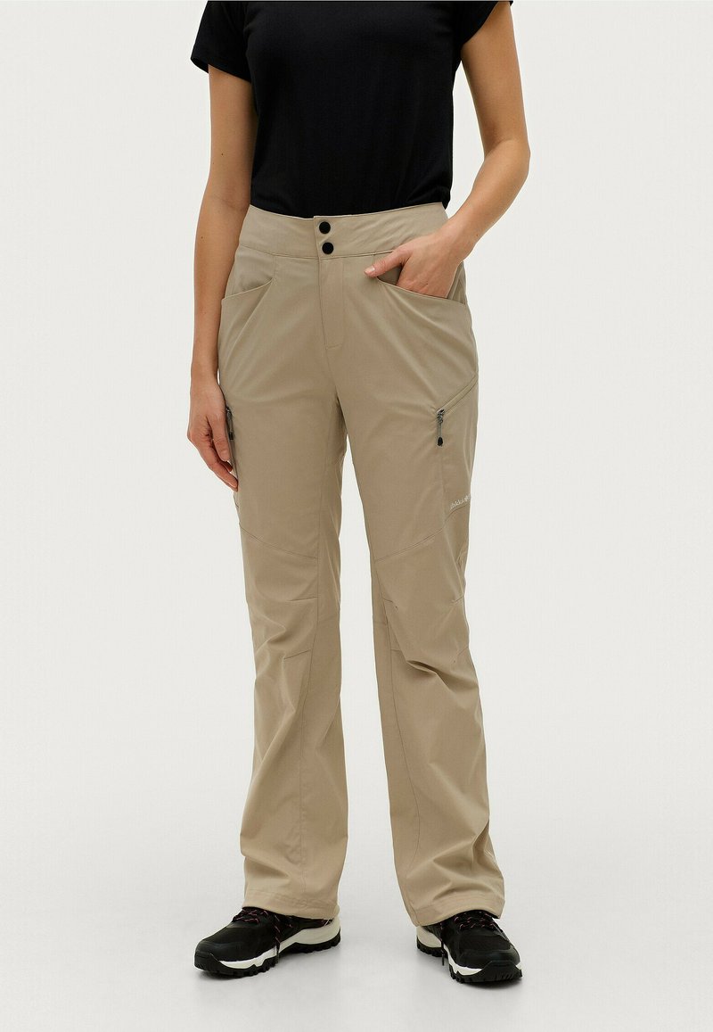 Beige outdoor broeken met een rechte pasvorm, voorzien van twee steekzakken en subtiele ritsen. Draagt een zwart shirt en zwarte schoenen.