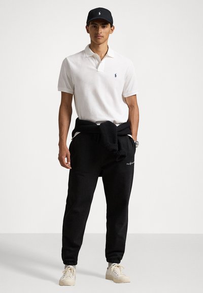 Weißes Polohemd, schwarze Jogginghose und weiße Turnschuhe. Das Polohemd hat einen Kragen und die Hose hat einen elastischen Bund mit Kordelzug. Schwarze Kappe getragen.