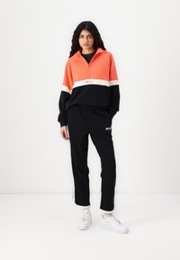 Felpa Roxy arancione e nera con strisce bianche; presenta un colletto con mezza zip. Pantaloni felpa neri abbinati con logo Roxy bianco. Sneakers bianche.