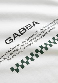 Λευκό ύφασμα με μαύρο κείμενο "GABBA" και μικρότερο κείμενο για το Neighbour Club, μαζί με πράσινο καρό μοτίβο κάτω από το κείμενο.