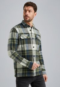 Chemise à carreaux en vert, marine et blanc. Dotée de deux poches poitrines, d'une fermeture à bouton et de manches longues. Tissu doux et texturé.