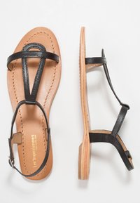 Sandali neri in pelle con una soletta color cuoio, caratterizzati da un design a T, minimal hardware e una suola piatta leggermente strutturata.