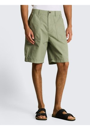 Mann trägt olivgrüne Cargo-Shorts und schwarze Sandalen mit Schnallen, steht vor einem einfarbigen hellen Hintergrund.