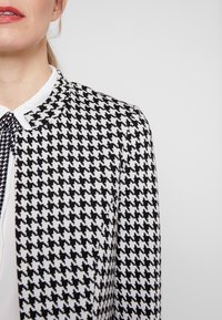 Blazer in bianco e nero a motivo houndstooth con colletto rotondo. Il tessuto presenta un motivo testurizzato con contrasti netti. Una camicetta bianca fa capolino da sotto.