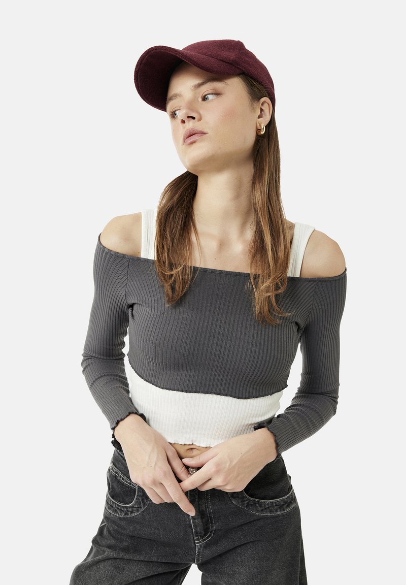 Joven mujer con gorra granate, top de canalé gris y blanco sin hombros, vaqueros oscuros, con pelo largo castaño, mirando hacia un lado sobre un fondo liso.