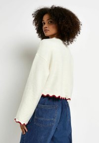 Krämfärgad croppad tröja med en vågig röd nederkant och ärmslut. Mjuk textur, avslappnad passform, i kombination med högmidjade mörkblå jeans.