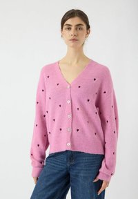 Rosa gestrickte Strickjacke mit V-Ausschnitt, verziert mit kleinen dunklen Herzmustern und weißen Knöpfen. Rippstruktur, Oversize-Schnitt und lange Ärmel.