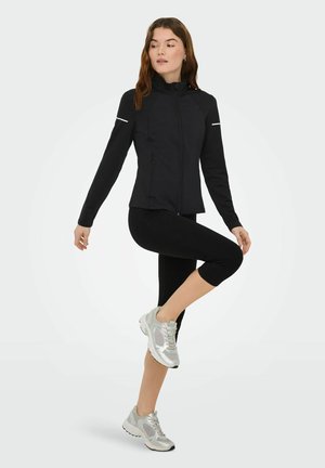 Melna sporta jaka ar rāvējslēdzēju priekšpusē, augstu apkakli un atstarojošām detaļām, kopā ar melnām kapri sporta biksēm un sudraba sporta apaviem.