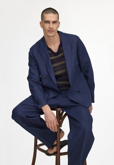 Modèle masculin assis sur un tabouret en bois, portant un costume bleu marine, un pull rayé bleu marine et beige, et des sandales noires, regardant droit devant.
