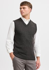 Mørkegrå ærmeløs sweater med V-hals, ribbet halsudskæring og kant, båret over en hvid langærmet skjorte, præsenteret mod en ensfarvet baggrund.