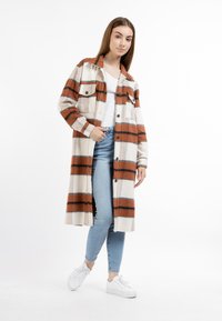 Giacca a camicia a quadri in crema e ruggine con tasche, chiusura a bottoni e vestibilità rilassata. Indossata sopra una maglietta bianca e pantaloni di jeans. Sneakers visibili.