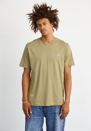 Camiseta de algodón verde oliva con cuello redondo y mangas cortas. Presenta un pequeño logo colorido en el lado izquierdo del pecho. Jeans de denim visibles por debajo.