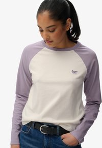 Camiseta de manga larga con un diseño de raglán lavanda, cuerpo en crema, textura suave de algodón, presenta un pequeño logo bordado en el pecho.