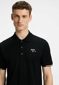 Czarna koszulka polo z trzema guzikami, z białym logo i kolorowymi akcentami. Krótkie rękawy z pasiastymi mankietami. Gładka faktura.