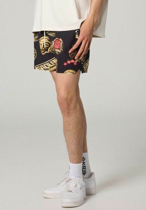 Male wearing black shorts with gold and red graphic designs, white graphic socks, white sneakers, and a white oversized T-shirt.