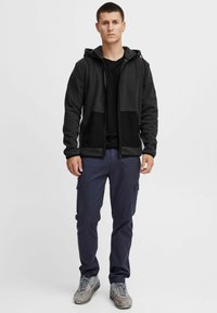 Sort fleece zip-up hoodie med kængurulommer, parret med marineblå cargo bukser. Modellen har grå sneakers på. Enkel, funktionel design.