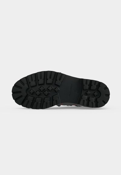Semelle de botte noire robuste avec des crampons profonds et des grips en forme de croix visibles sur un fond clair uni.
