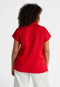 Camicia rossa a maniche corte realizzata in tessuto leggero, caratterizzata da un colletto a piatto e una texture liscia, con un orlo arrotondato e un dettagliato spacco posteriore.