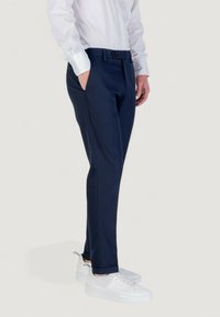 Homme portant un pantalon ajusté bleu marine, une chemise blanche et des baskets blanches, se tenant avec une main dans sa poche contre un fond uni.